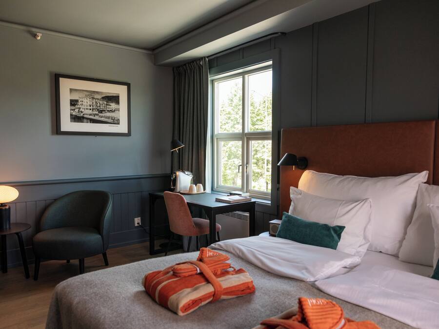 Une chambre d’hôtel avec un lit, un bureau, une chaise, une lampe et une fenêtre avec des rideaux.