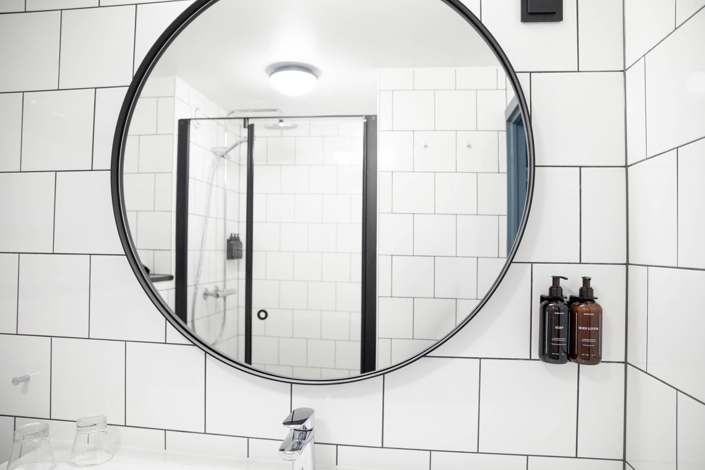 A bathroom with white tiled walls, a round mirror, a shower, and two bottles of soap on the wall.