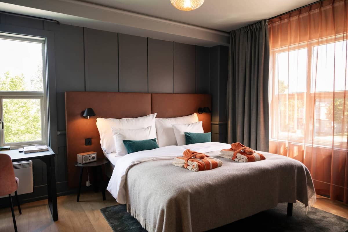 A hotel room with a large bed, a desk, a chair, a small table with a radio, and a window with curtains.