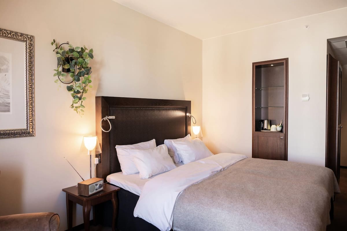A hotel room with a bed, bedside lamps, a small table, a plant, a framed picture, and a wardrobe.