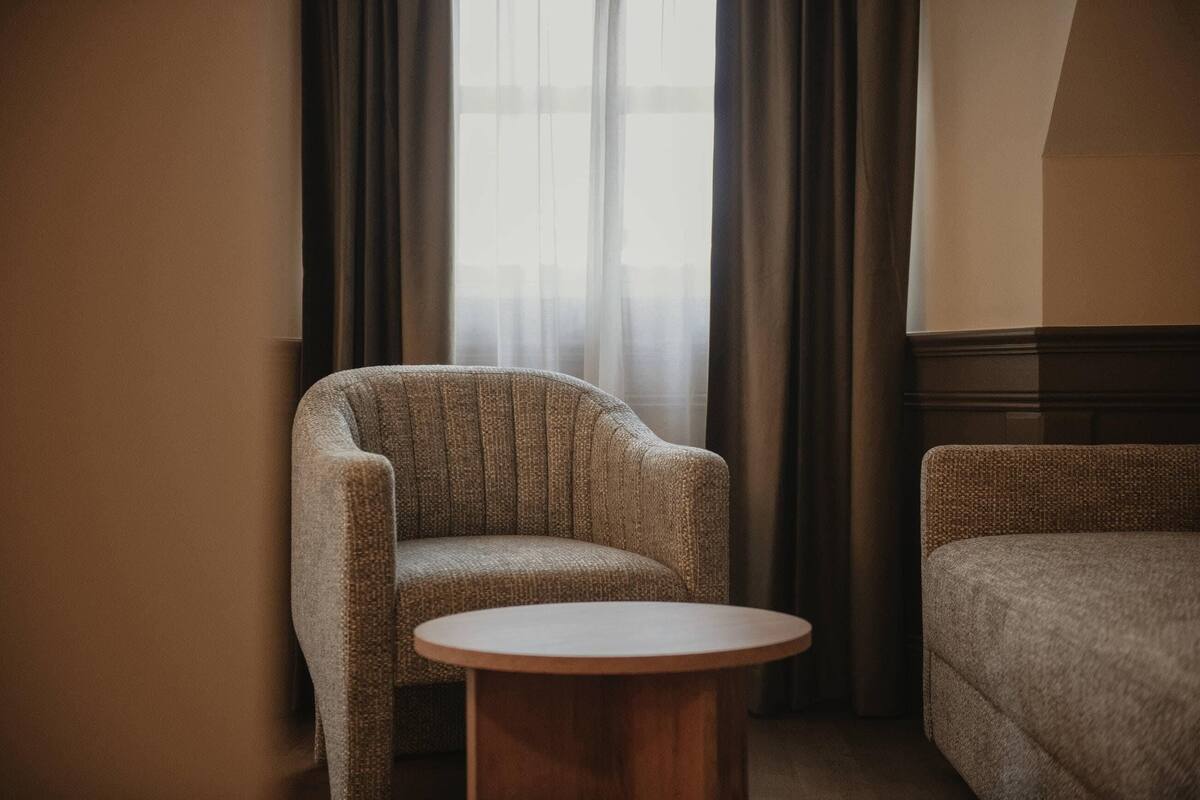A room with a single armchair, a round wooden table, and a window with sheer curtains.