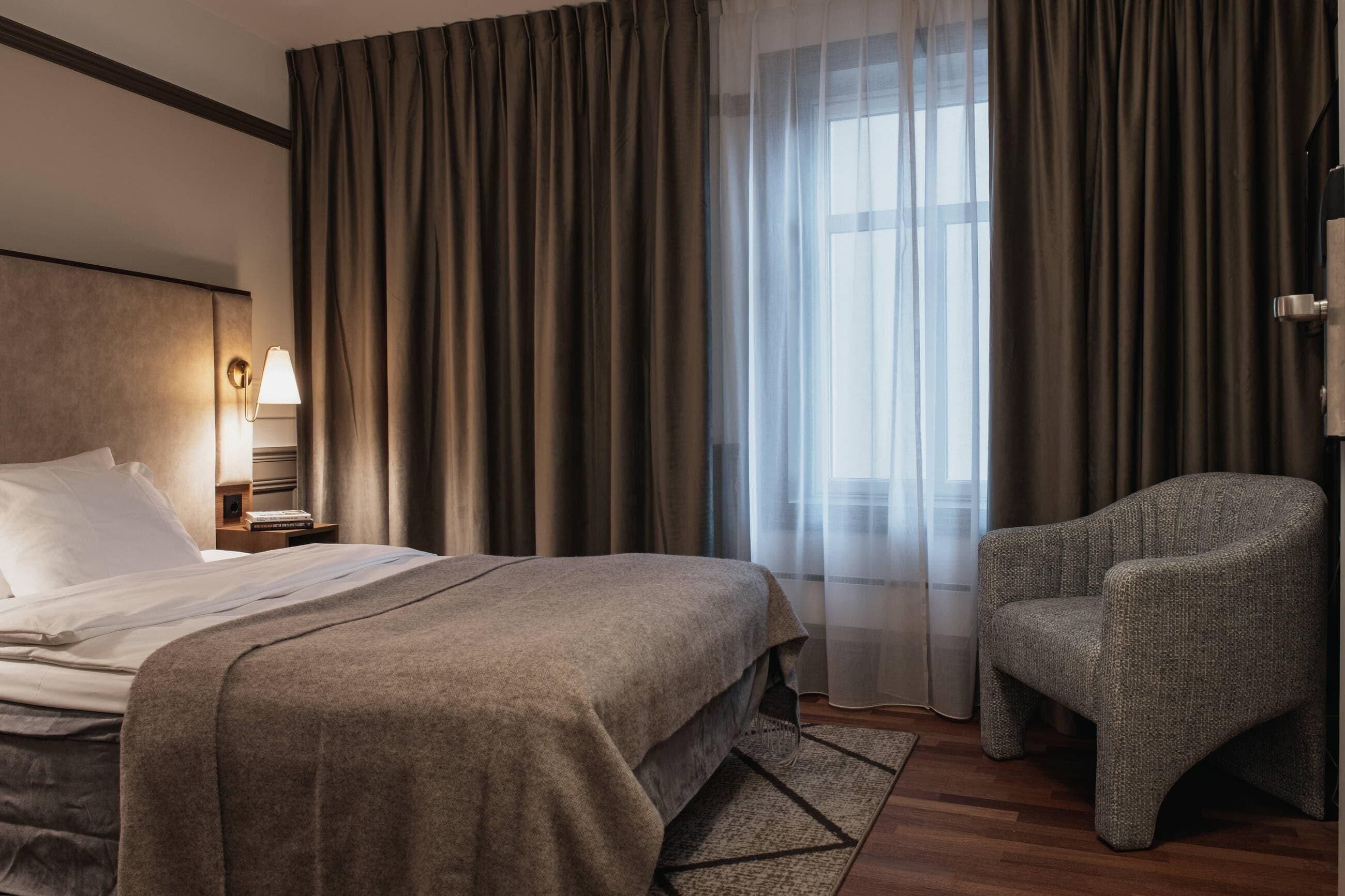 Une chambre d’hôtel avec un lit, une chaise, une table de chevet avec une lampe et une fenêtre avec des rideaux.