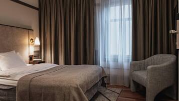 Une chambre d’hôtel avec un lit, une chaise, une table de chevet avec une lampe et une fenêtre avec des rideaux.