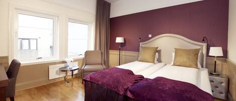 A hotel room with a bed, two bedside lamps, a chair, a small table, and a window with curtains.
