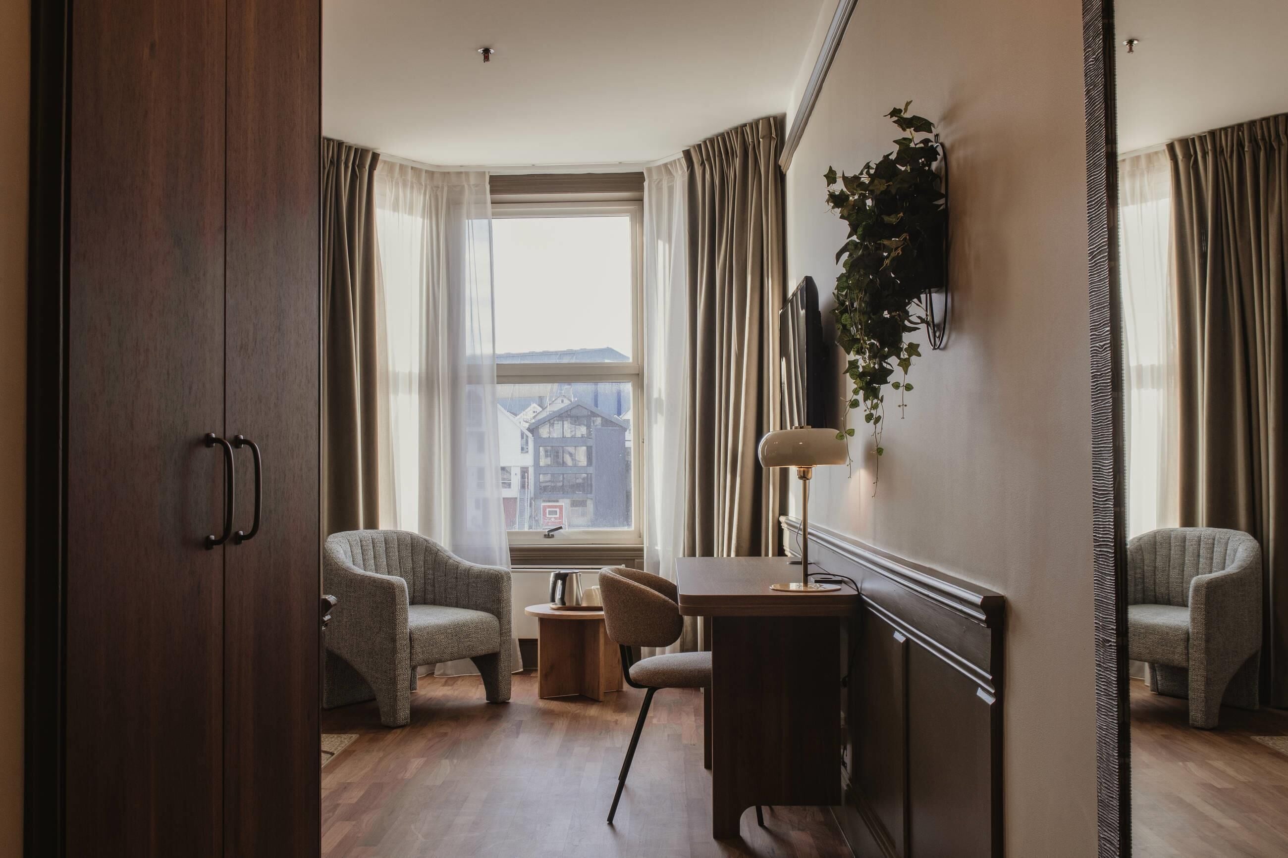 A room with a wooden wardrobe, two armchairs, a desk with a lamp, and a view of buildings through a window with curtains.