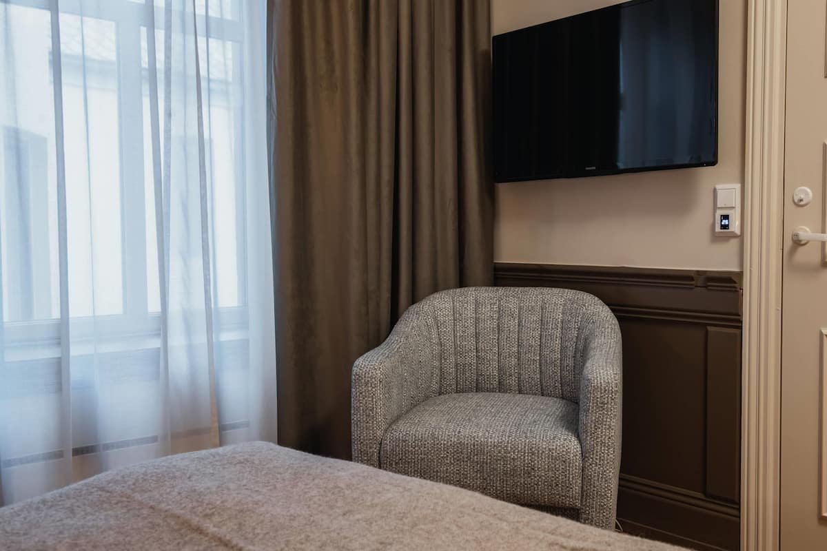 A hotel room with a bed, a chair, a television mounted on the wall, and a window with curtains.