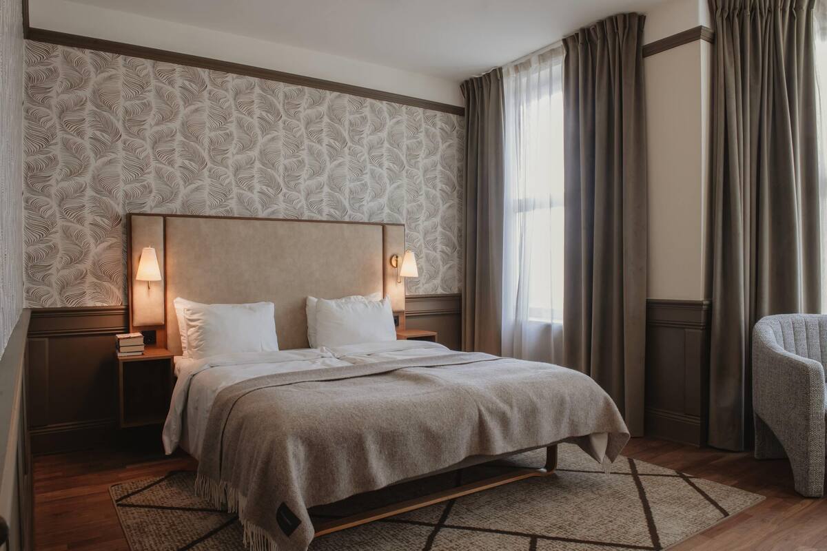 A hotel room with a large bed, two bedside lamps, a nightstand with books, a patterned wallpaper, and a view of the outdoors.