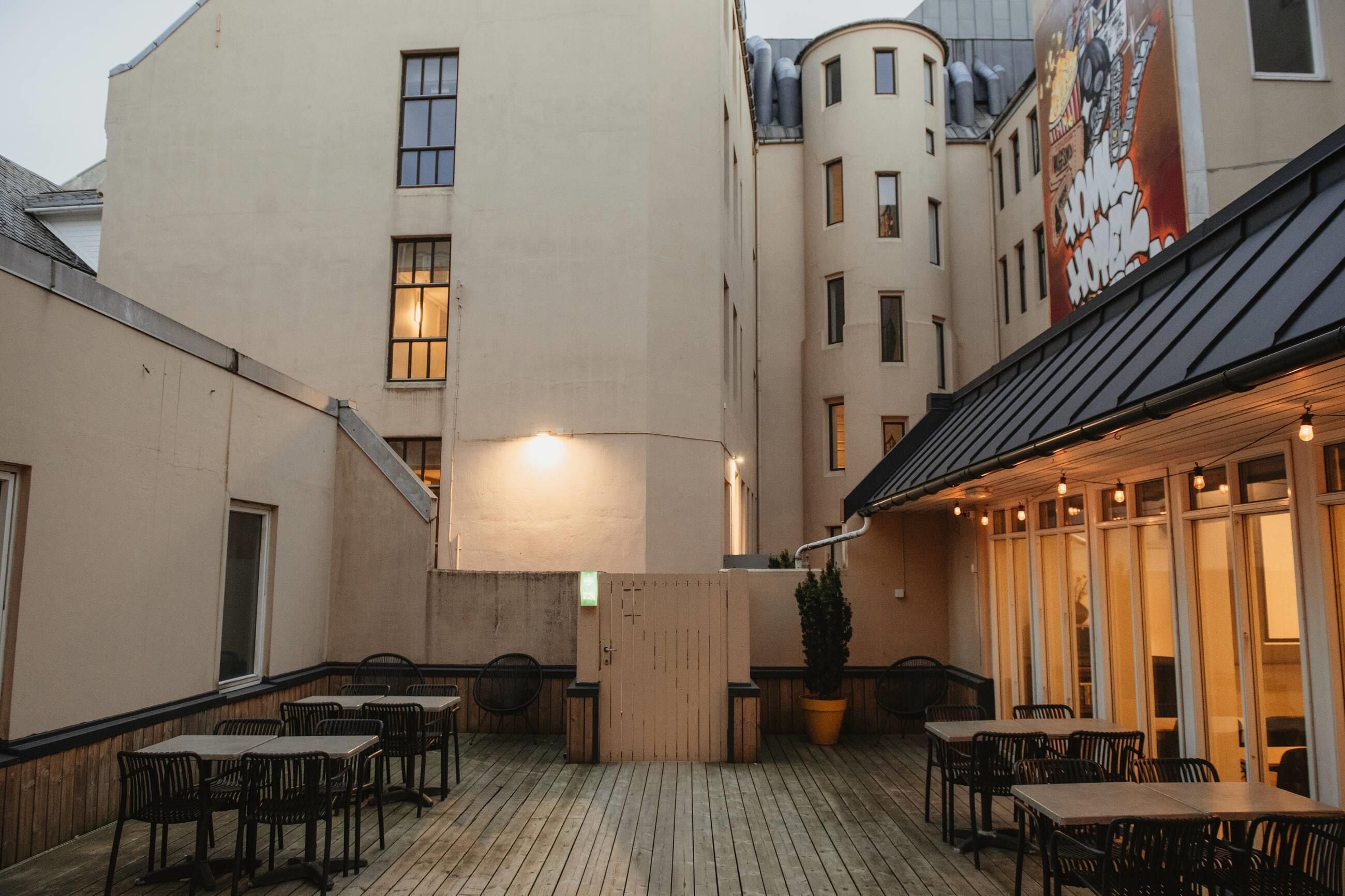 An outdoor patio area with tables and chairs, a wooden deck, and a building with large windows and a mural.
