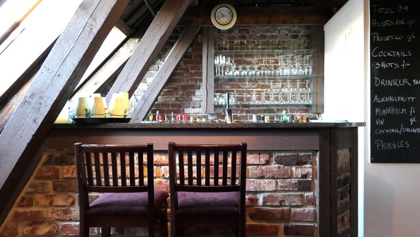 A bar area with wooden chairs, a brick wall, and a chalkboard menu listing drinks and shots. - Home Hotel Amanda (Haugesund)