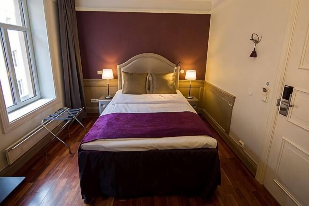 A hotel room with a bed, two bedside lamps, a window with curtains, a radiator, and a door.