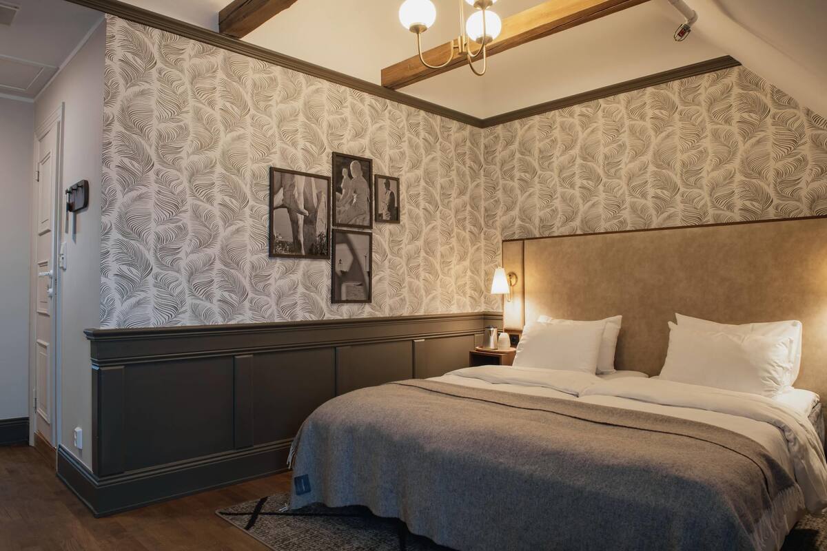 A bedroom with a patterned wallpaper, a bed with white linens, a nightstand with a lamp, and a framed picture on the wall.