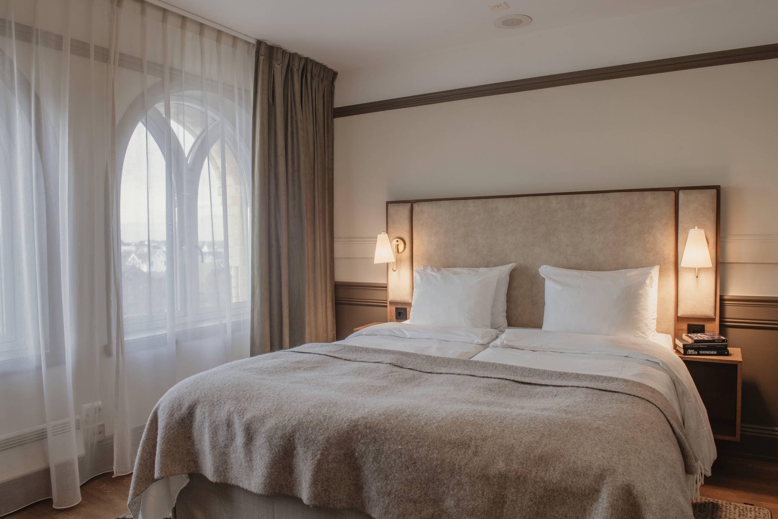 Ein Hotelzimmer mit einem großen Bett, zwei Nachttischlampen, einem Nachttisch mit einem Buch und einem Fenster mit durchsichtigen Vorhängen.