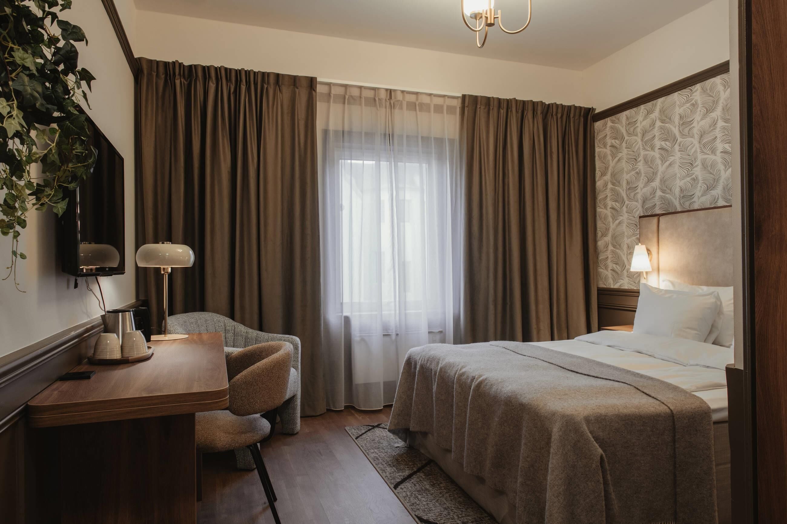A hotel room with a bed, a desk with a lamp, a chair, a large window with curtains, and a wall-mounted TV.