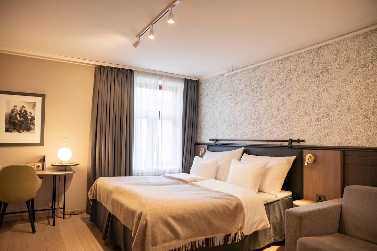 A hotel room with a bed, bedside table, lamp, chair, and a framed picture on the wall.