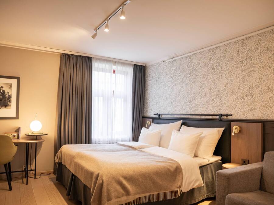 A hotel room with a bed, bedside table, lamp, chair, and a framed picture on the wall.