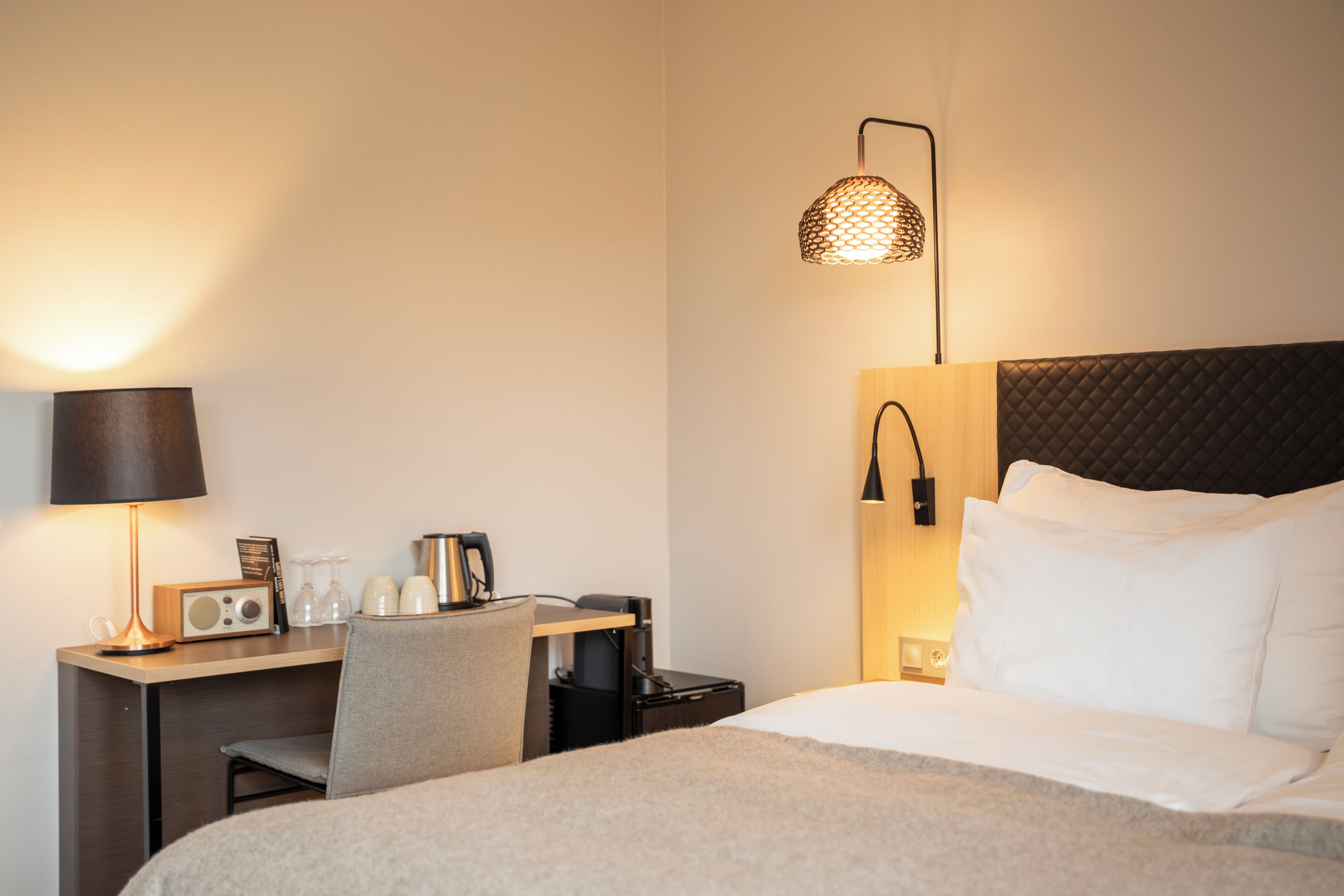 A hotel room with a bed, a desk with a lamp, a radio, and a wall-mounted lamp.