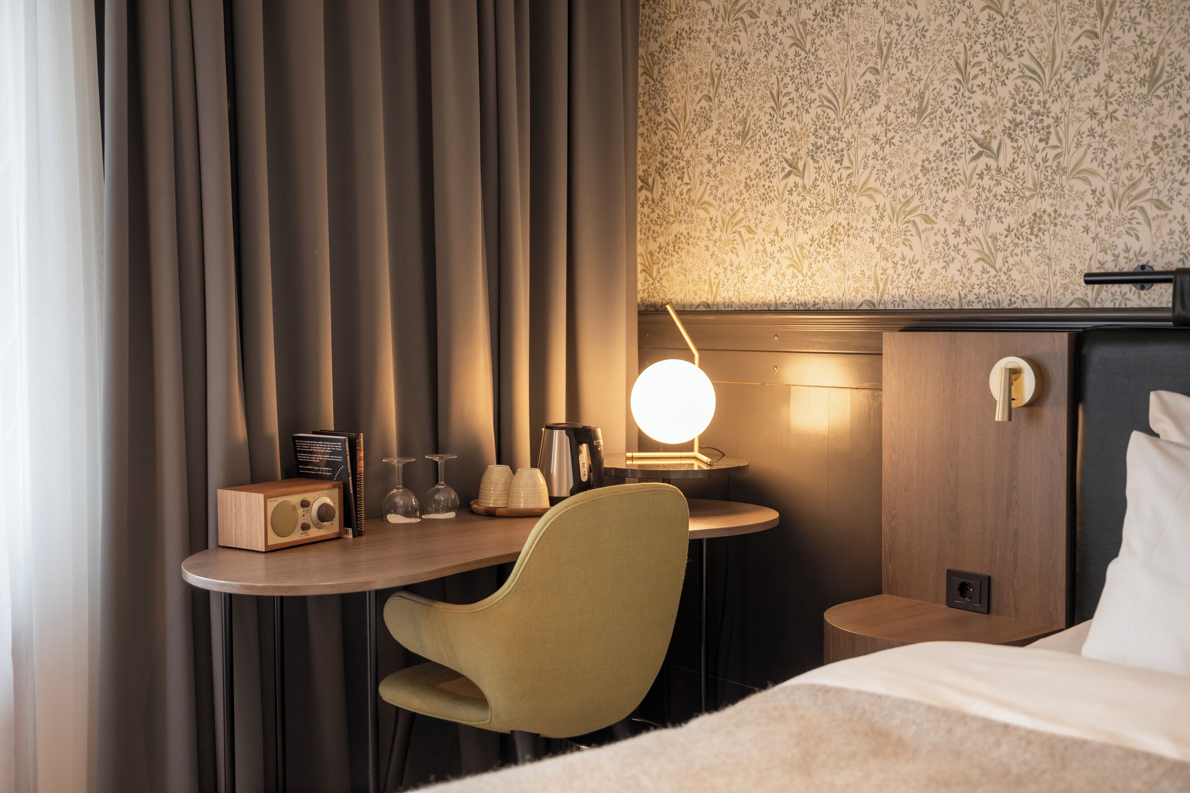 A hotel room with a bed, a desk with a lamp, a chair, and a small table with a radio and glasses.