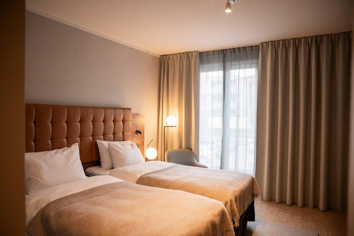 A hotel room with a large bed, a smaller bed, a nightstand with a lamp, a chair, and a window with curtains.