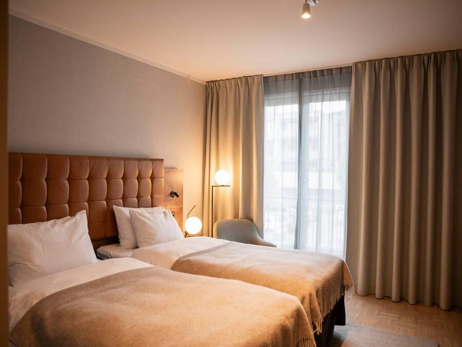 A hotel room with a large bed, a smaller bed, a nightstand with a lamp, a chair, and a window with curtains.