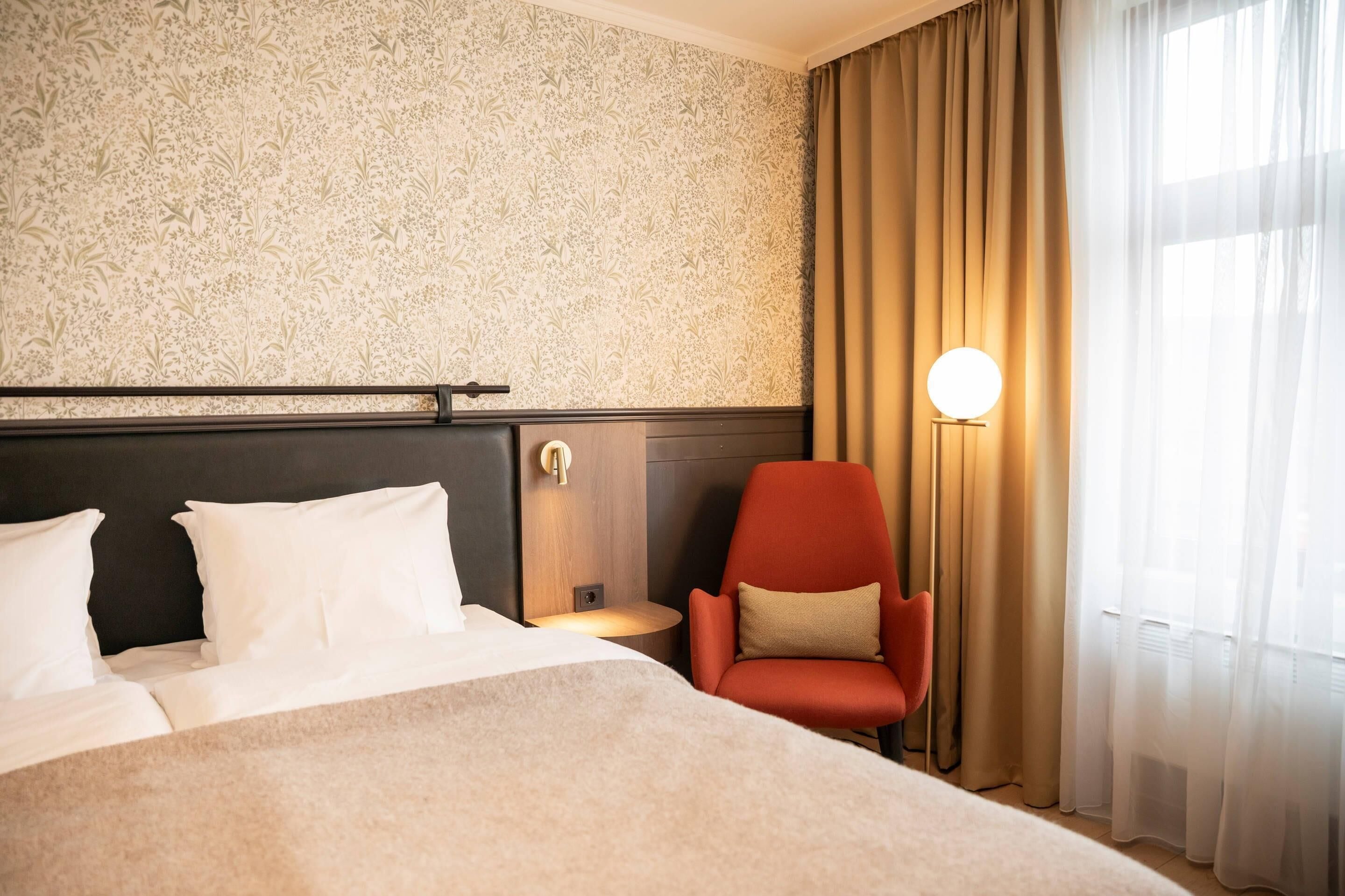 Habitación de hotel con una cama grande, una silla roja, una lámpara fijada a la pared y un ventanal con cortinas.