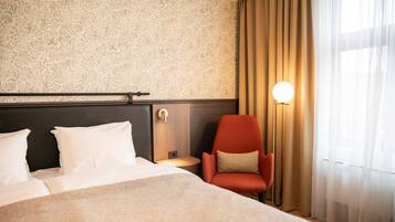 Habitación de hotel con una cama grande, una silla roja, una lámpara fijada a la pared y un ventanal con cortinas.