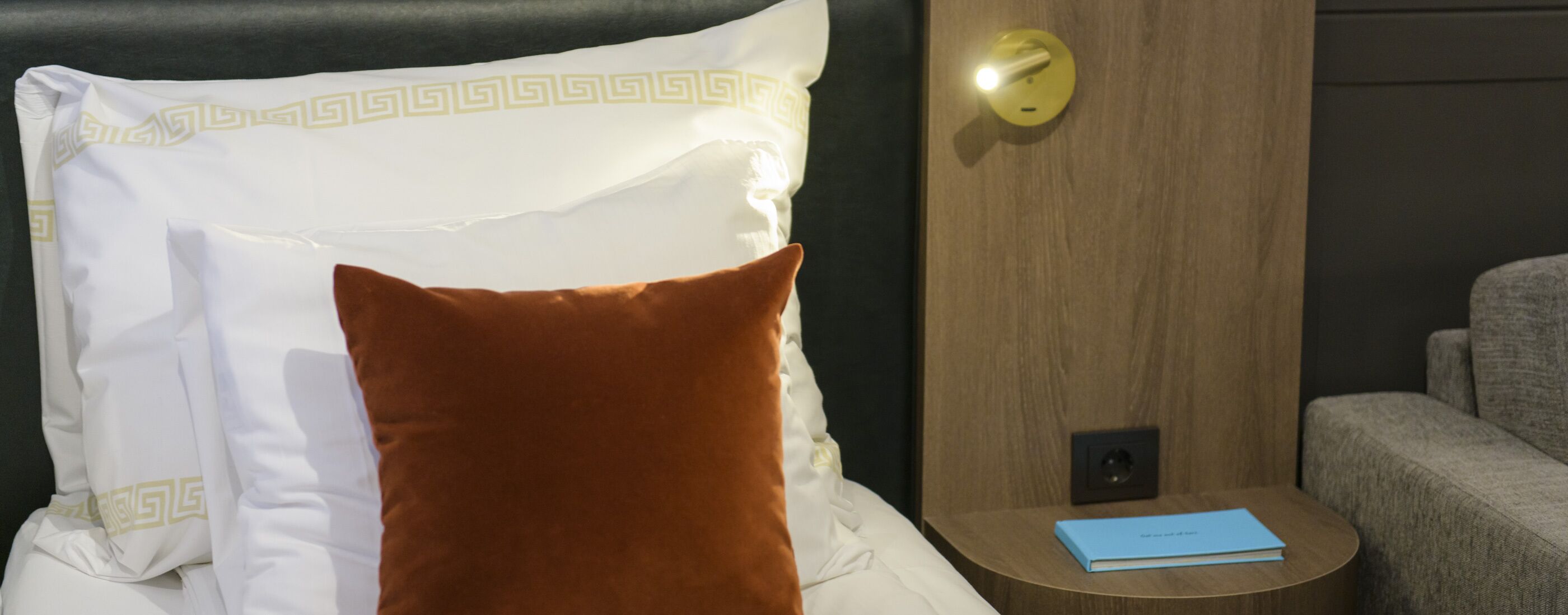 Una cama bien hecha con un almohadón blanco de diseño geométrico, un almohadón marrón y un libro azul sobre una mesita de noche de madera.