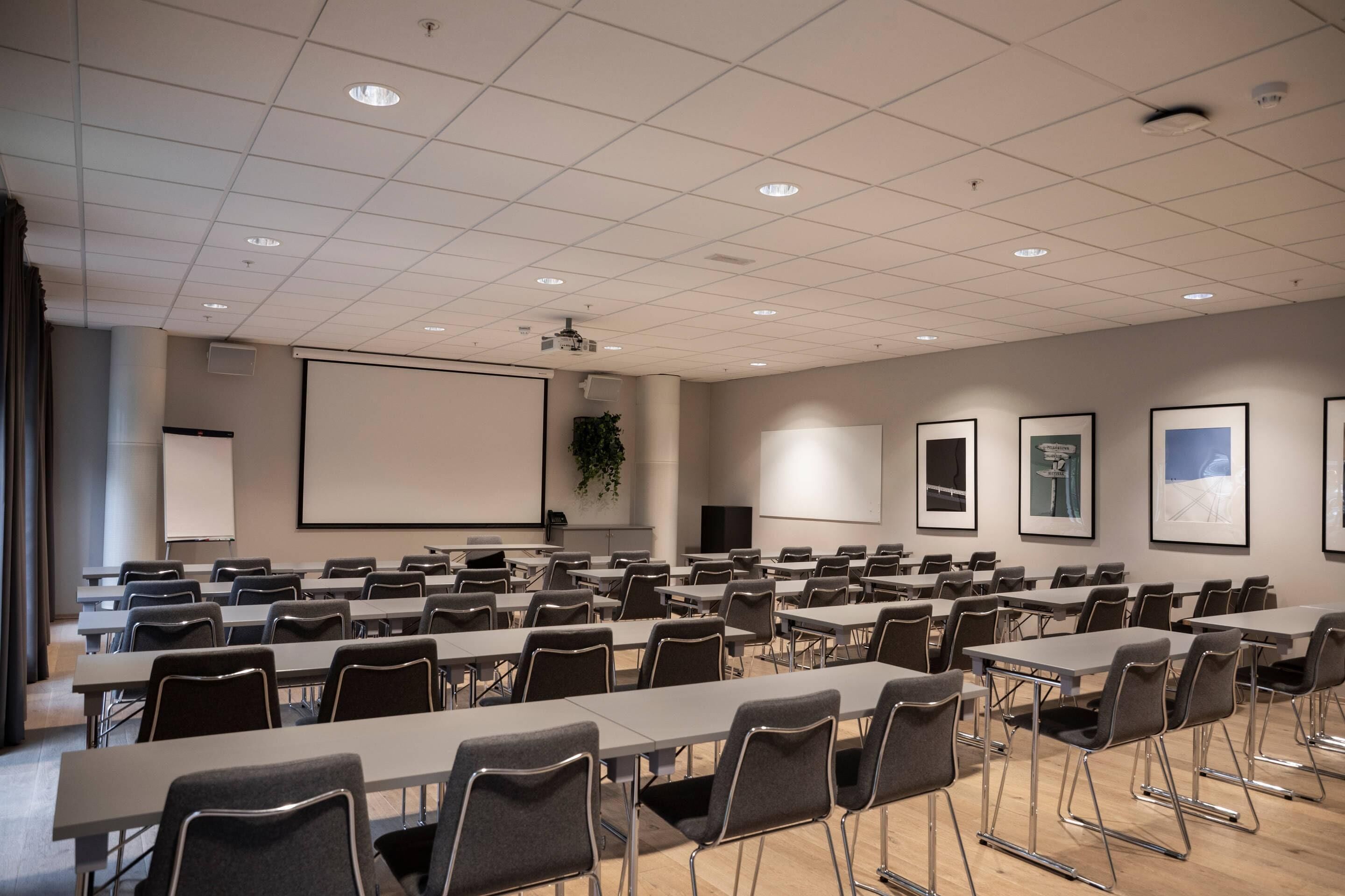 Ein Konferenzraum mit Tischen und Stühlen, einer Projektionsleinwand und gerahmten Bildern an der Wand.