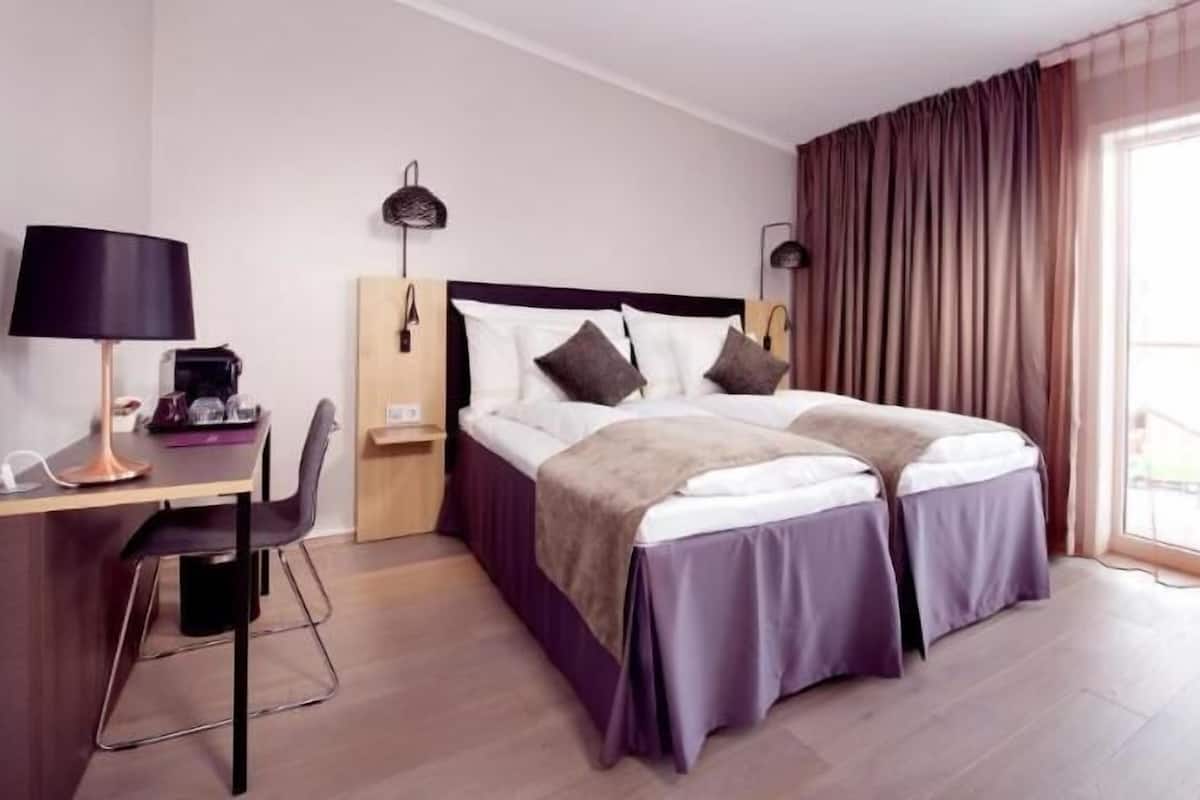 A hotel room with a bed, two bedside tables, a desk with a chair, a lamp, and a window with curtains.