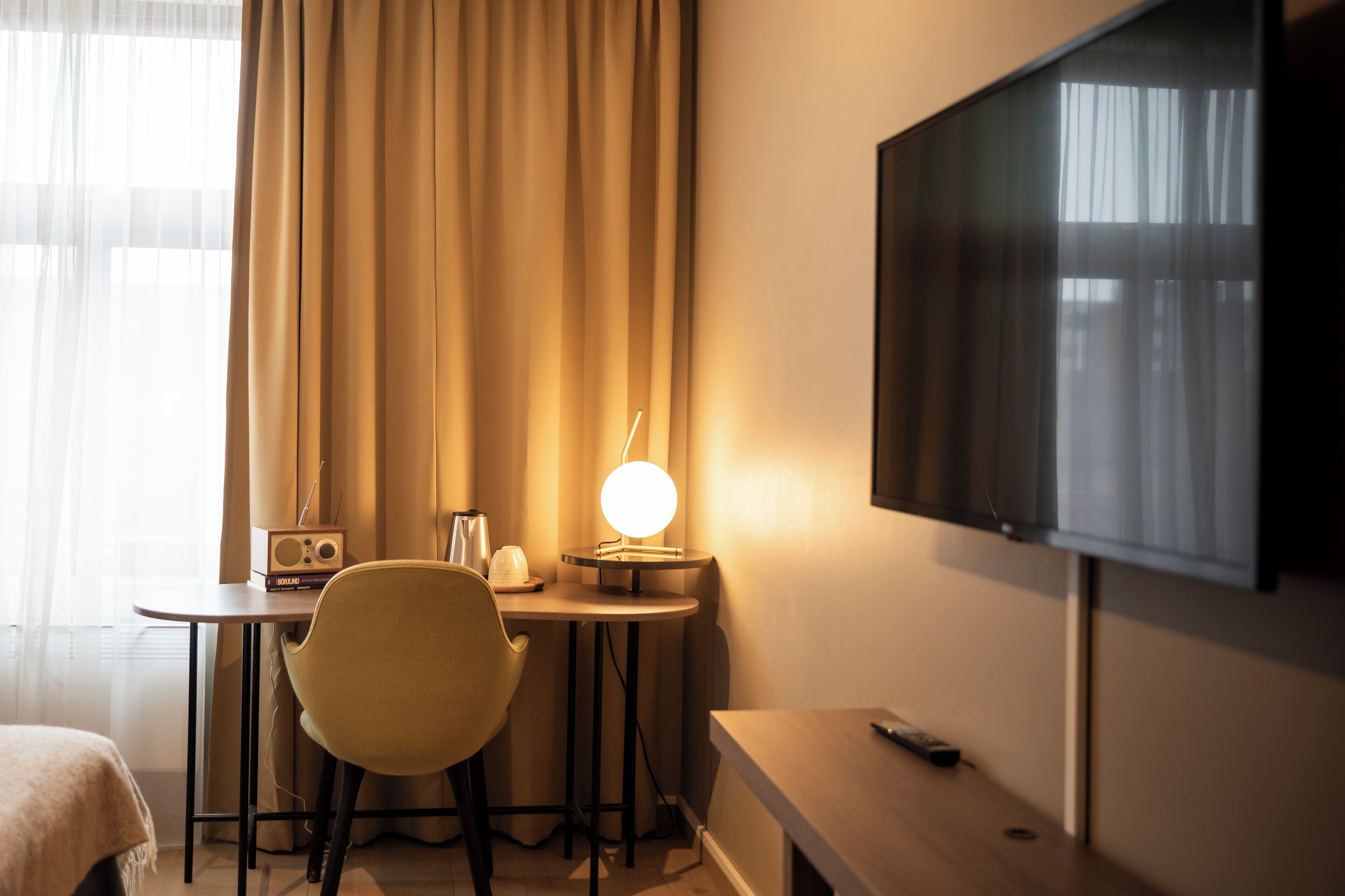 A hotel room with a bed, a desk with a lamp, a chair, a television, and a window with curtains.