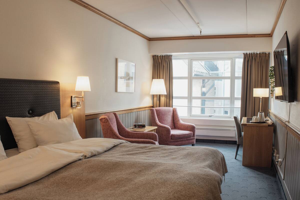 Une chambre d’hôtel avec un grand lit, deux fauteuils, un bureau, une télévision et une fenêtre avec des rideaux.