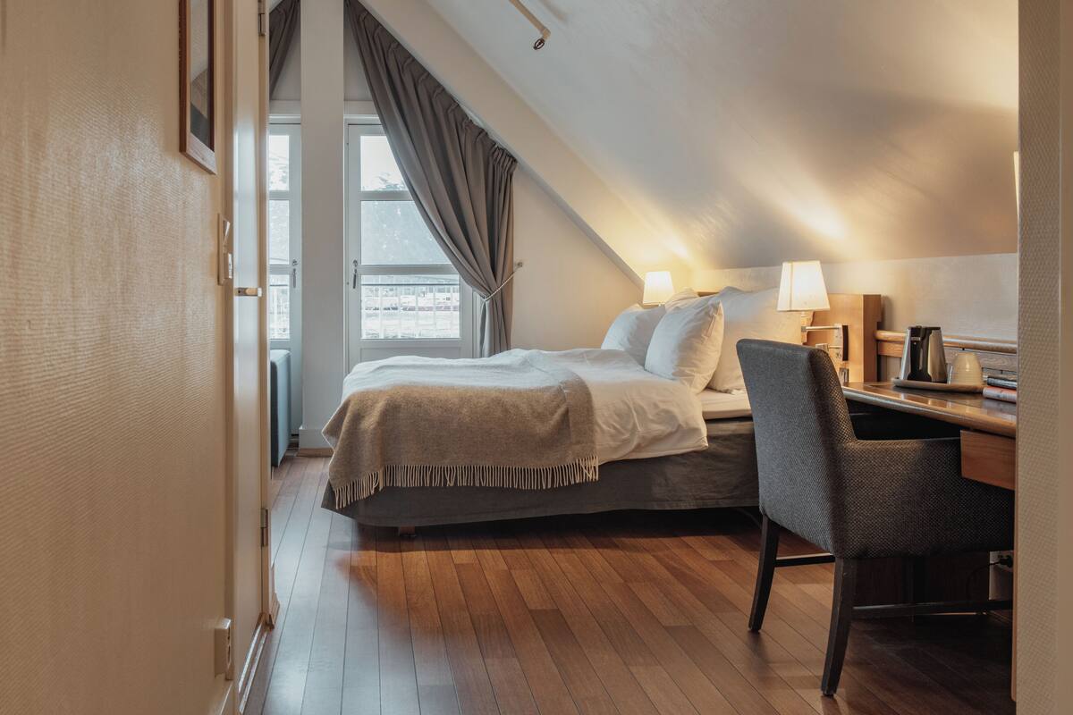 Une chambre à coucher avec un lit, un bureau, une chaise, une lampe et une fenêtre avec des rideaux.