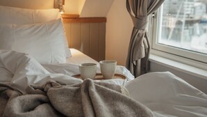 Een hotelkamer met een bed, kussens, een nachtkastje met een lamp en een raam met gordijnen.