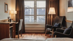 Een hotelkamer met een bureau, twee stoelen, een bed, een raam met gordijnen en een lamp.