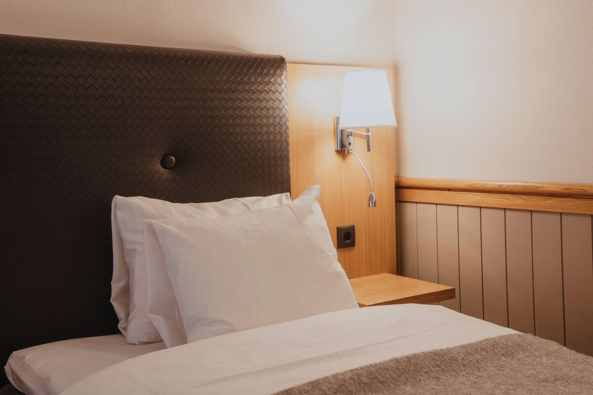 Un lit bien fait, avec du linge de lit blanc et des oreillers, une tête de lit en bois, une table de chevet avec une lampe et un luminaire fixé au mur.