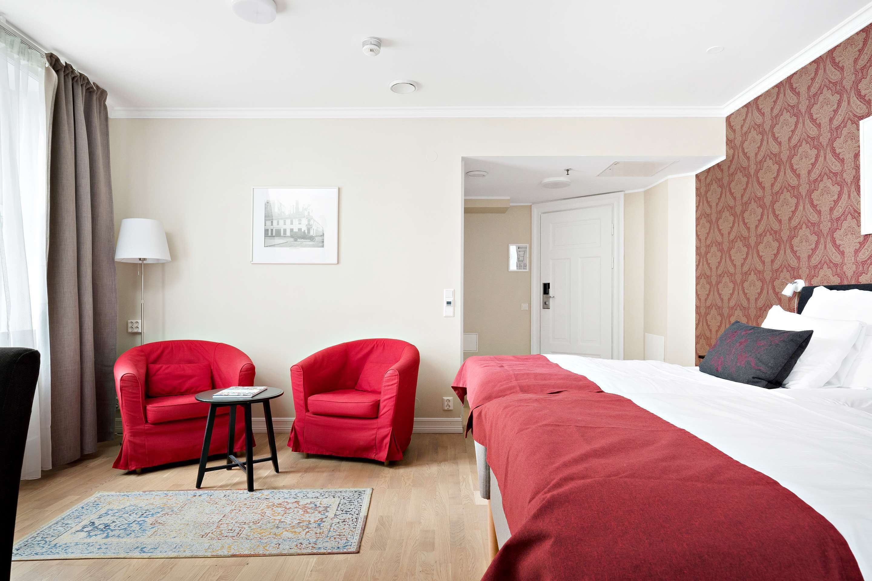 A hotel room with a bed, two red armchairs, a small table, a rug, and a door leading to another room.
