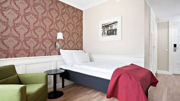 Ein Schlafzimmer mit einem Bett, einem grünen Sessel, einem kleinen Tisch, einer roten Tagesdecke, einem gemusterten Wandpapier und einem gerahmten Foto an der Wand.
