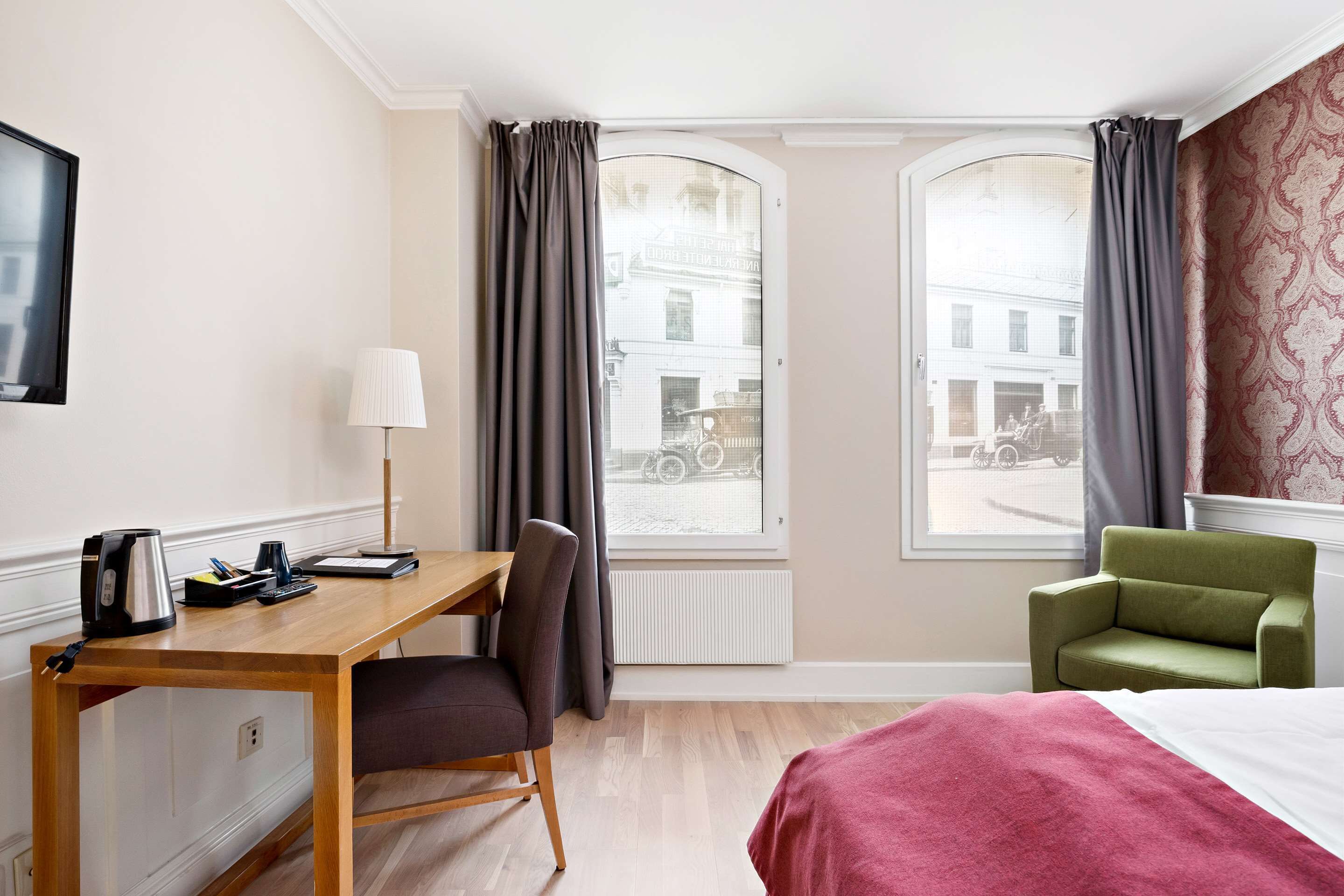 Quarto de hotel com escrivaninha de madeira, uma cadeira, cama com manta vermelha, poltrona verde e duas janelas com cortinas.