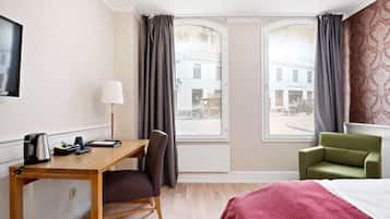 Quarto de hotel com escrivaninha de madeira, uma cadeira, cama com manta vermelha, poltrona verde e duas janelas com cortinas.