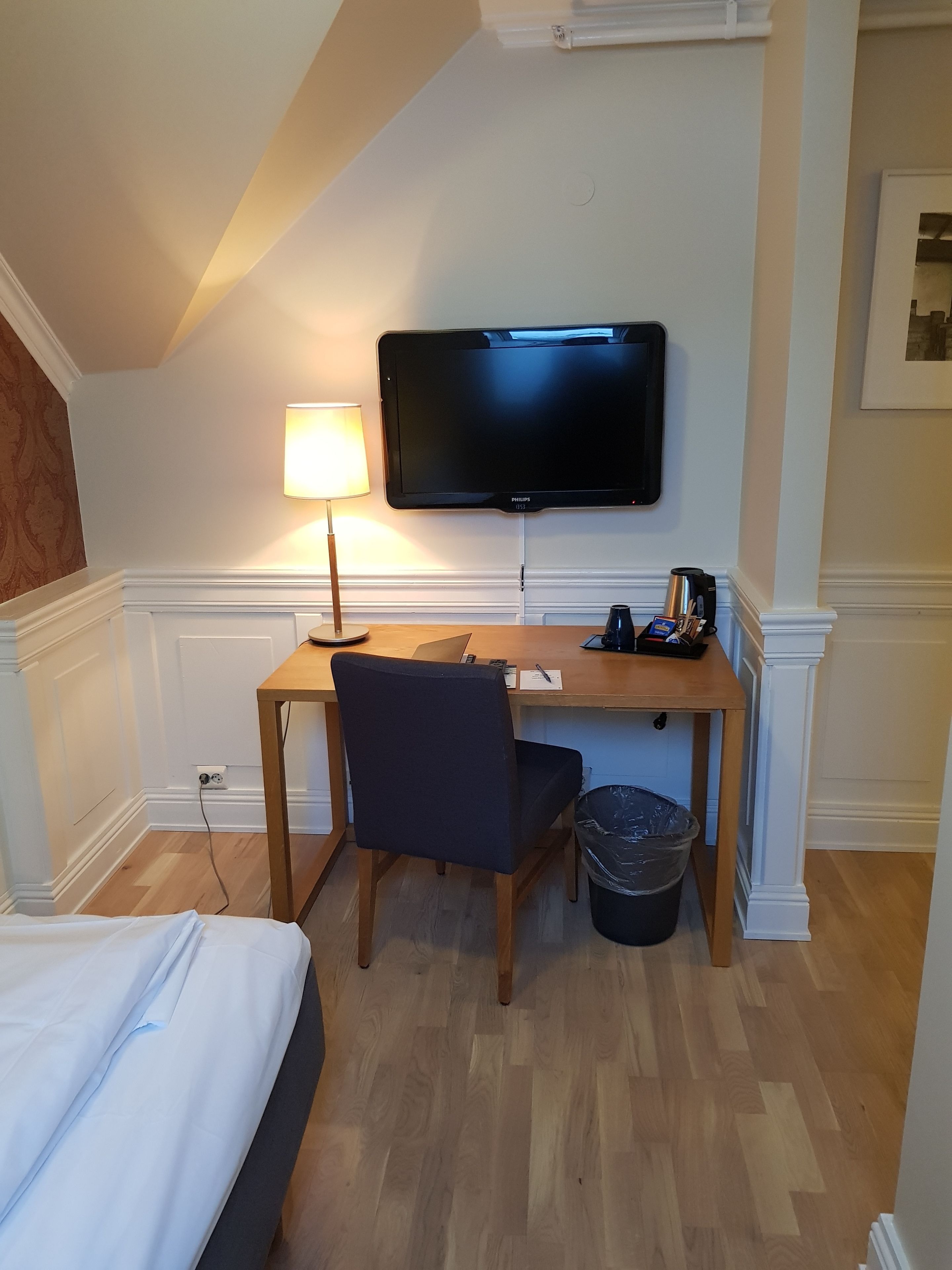 A hotel room with a bed, a desk with a chair, a television, a lamp, and a trash bin.