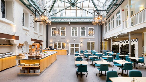 A spacious indoor area with a central serving counter, multiple tables and chairs, and a high ceiling with a glass structure. - Best Western Plus Hotel Bakeriet (Trondheim)