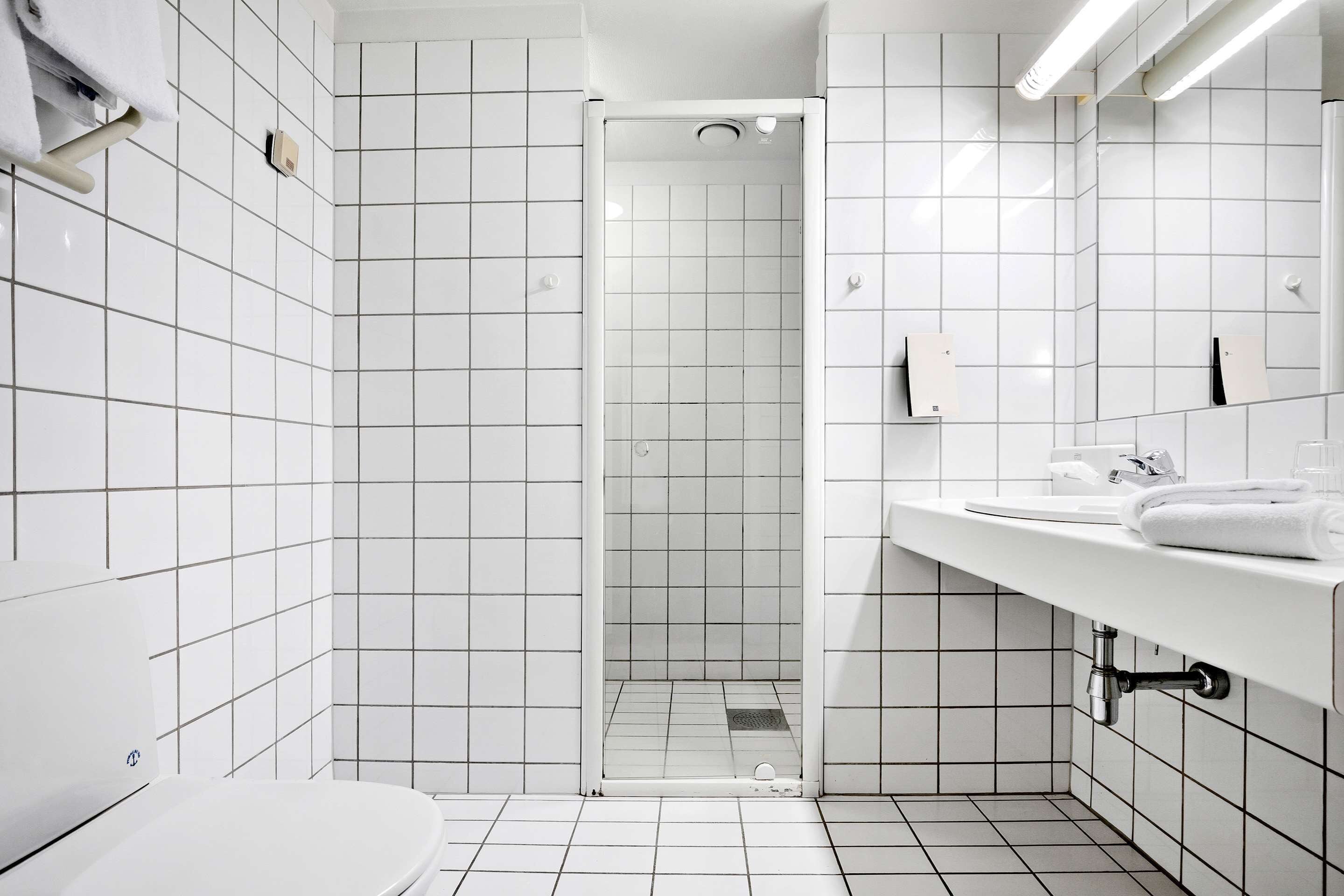 Un baño moderno con baldosas blancas, ducha con mampara de vidrio, lavamanos blanco y retrete.