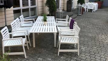 Et udendørs siddeområde med hvide borde og stole, omgivet af plantekrukker og en brobelagt gulv.