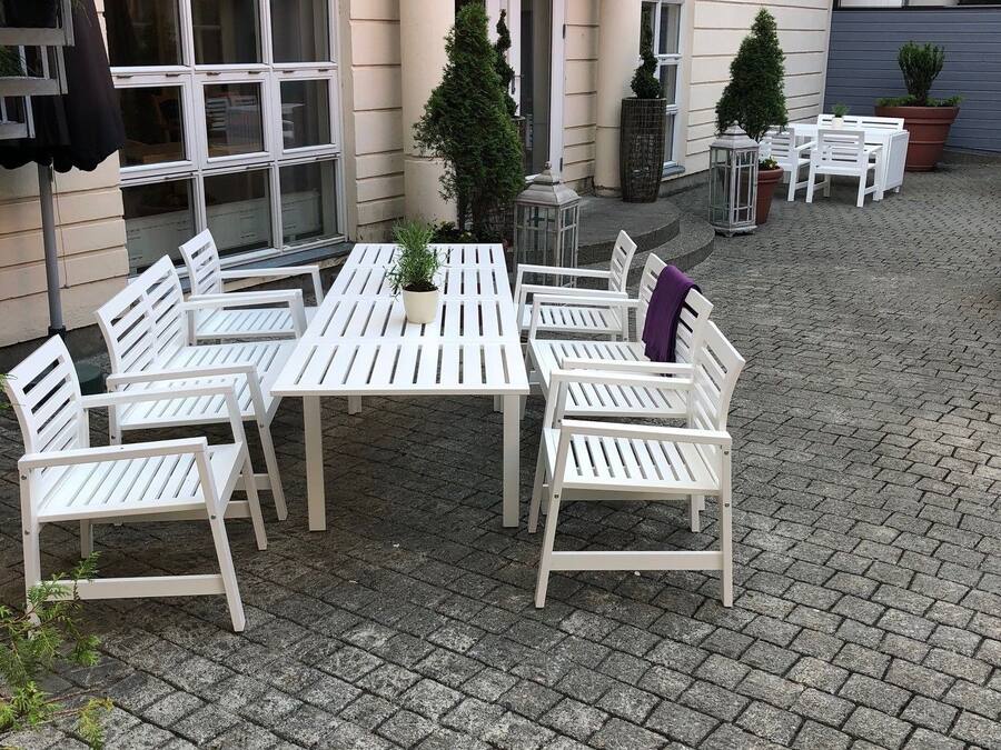 Et udendørs siddeområde med hvide borde og stole, omgivet af plantekrukker og en brobelagt gulv.