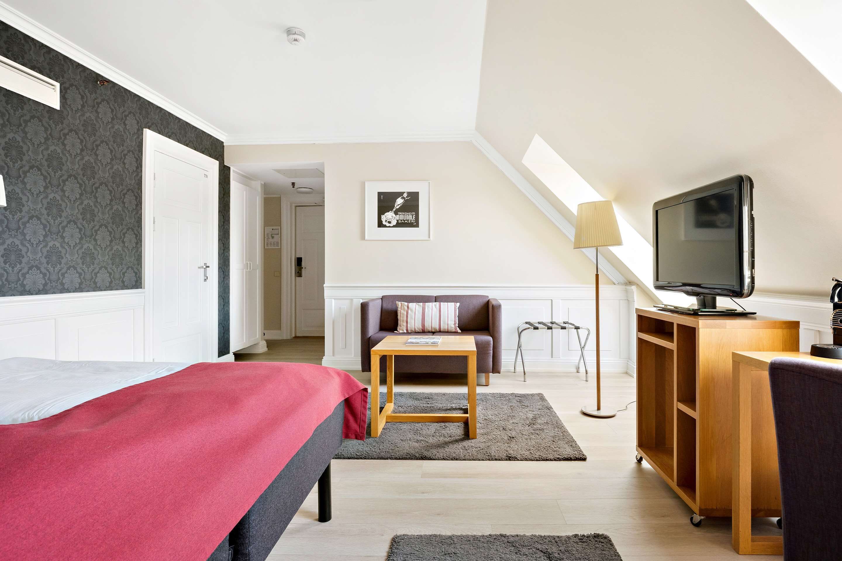A bedroom with a bed, a wooden nightstand, a TV, a desk, a sofa, and a lamp.