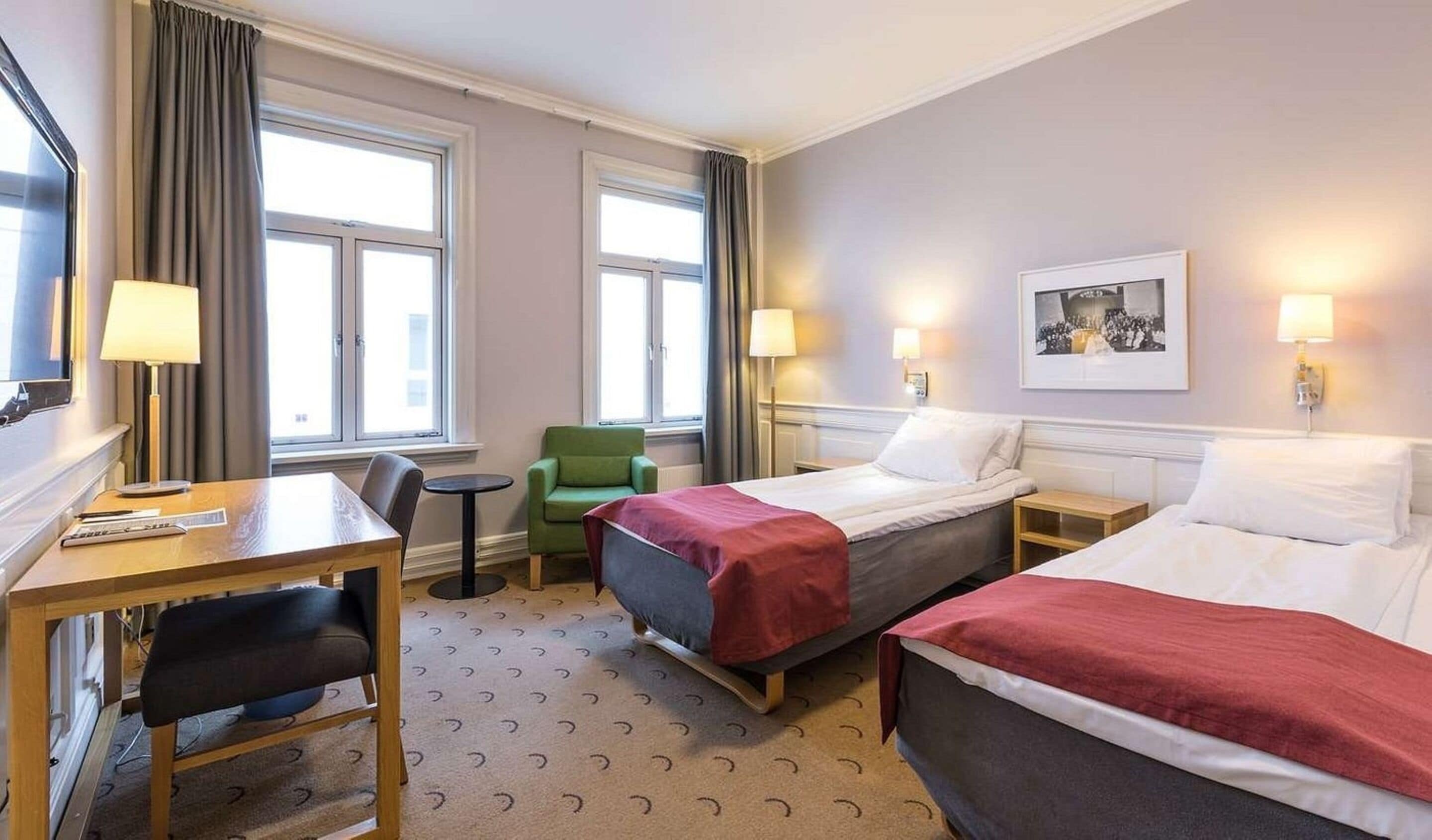 A hotel room with two beds, a desk, a chair, a small table, and two lamps.