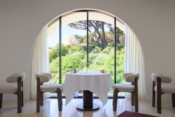 A dining area with a round table, four chairs, and a view through a large arched window.