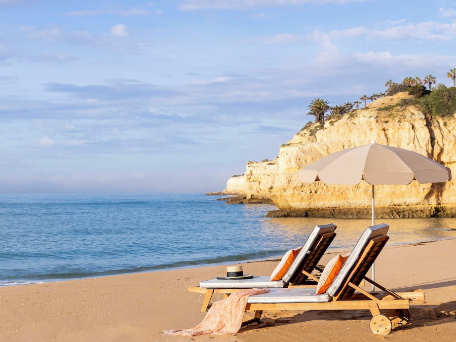A beach with two sun loungers under a beige umbrella, a rocky cliff in the background, and calm sea.