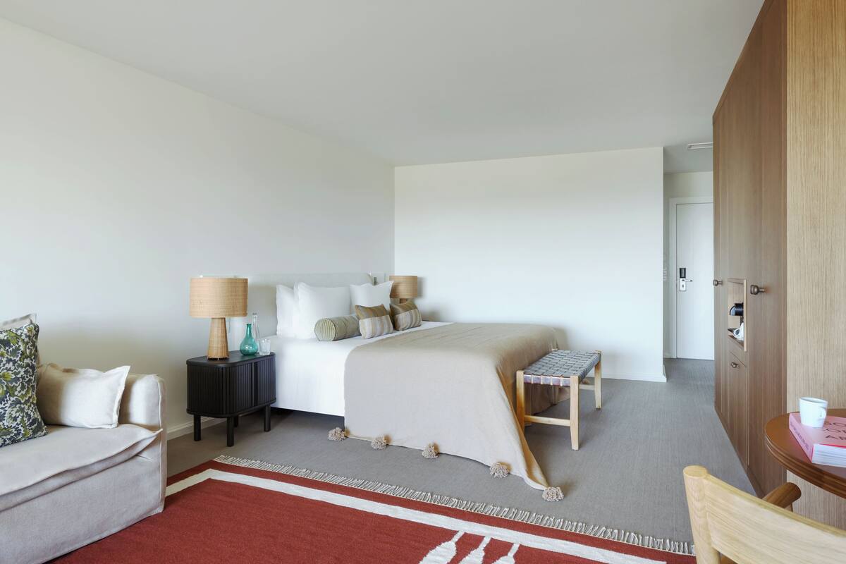 A hotel room with a bed, bedside tables, a chair, a small table, and a door.