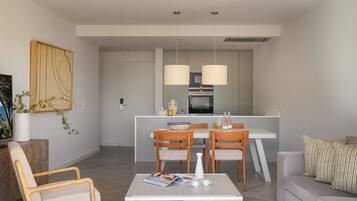 Una sala de estar moderna con un sofá, una mesa de centro y dos sillas de madera. Hay un comedor con una mesa y sillas, y una cocina al fondo.