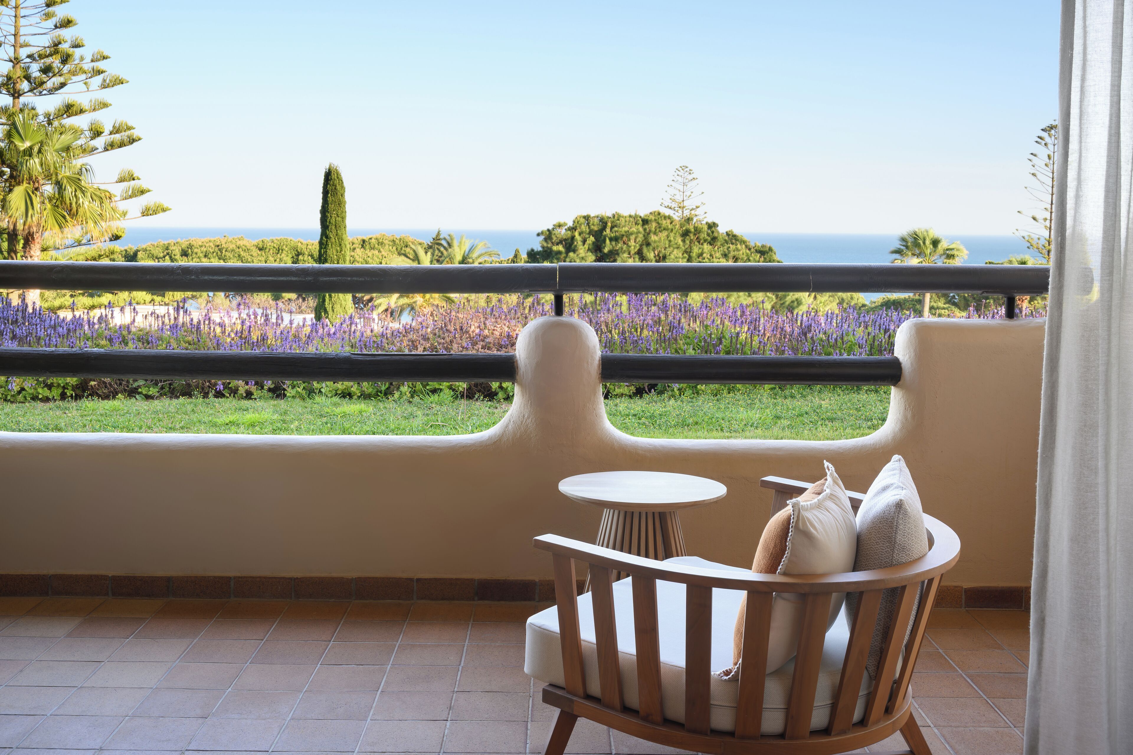 A balcony with a view of the sea, a chair with cushions, and a small table.